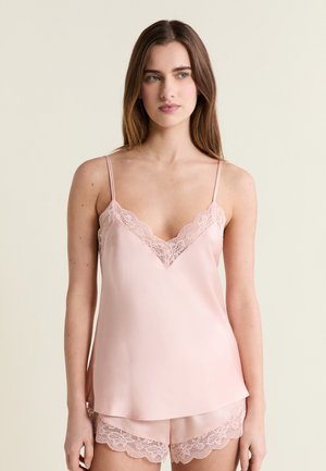 Kobieta ubrana w bladoróżową satynową koszulkę na ramiączkach i pasujące krótkie spodenki z koronkową lamówką, stojąca na tle jednolitego jasnego tła.