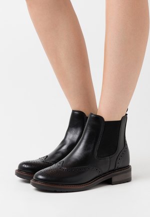 Botas de tobillo de cuero negro con detalles de brogue, llevadas sobre piernas desnudas, con paneles laterales elásticos y suelas con bordes marrones sobre un fondo blanco.