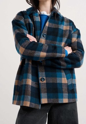 Personne portant une veste à carreaux bleue, noire et beige avec des boutons, les bras croisés sur un pull bleu et un pantalon noir sur un fond uni.