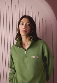 Green zip-up sweatshirt with a high collar and "PARIS" printed in pink. Soft fabric, relaxed fit, set against a pink textured background.