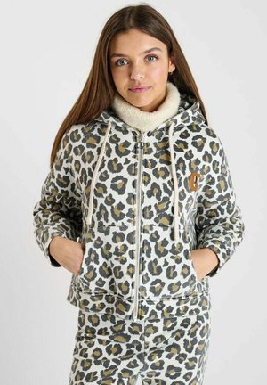 Jeune femme aux longs cheveux bruns portant un sweat à capuche zippé imprimé léopard et un pantalon assorti, les mains dans les poches.