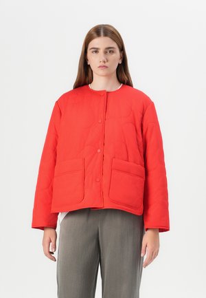 Model stands in a red quilted jacket with two front pockets, paired with gray trousers, against a plain white background.