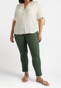 Vit, knäppt blus med korta, rullade ärmar och knytdetaljer, tillsammans med figurnära gröna byxor och beige sandaler.