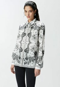 Camicia bianca con bottoni, caratterizzata da motivi floreali e geometrici neri, maniche lunghe e colletto classico, realizzata in un tessuto leggero.