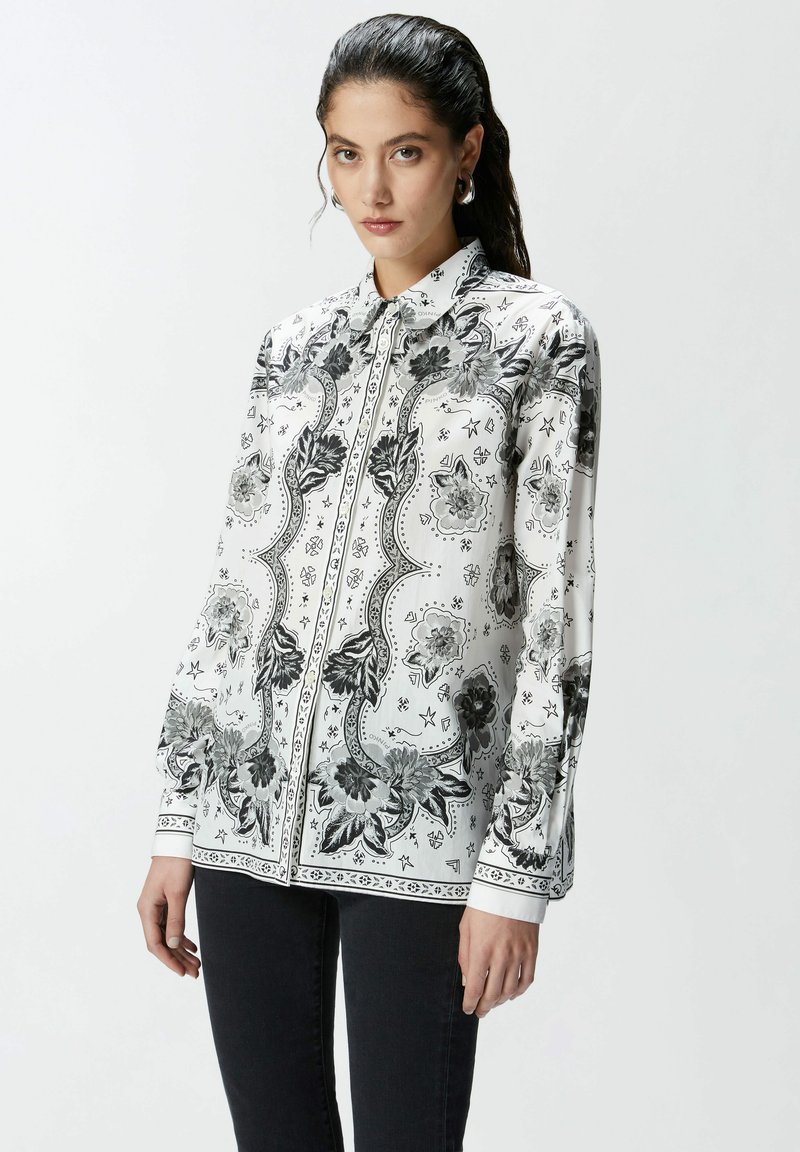 Camicia bianca con bottoni, caratterizzata da motivi floreali e geometrici neri, maniche lunghe e colletto classico, realizzata in un tessuto leggero.