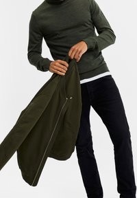 Olivgrön zip-up jacka som hålls i handen, tillverkad av slätt tyg; kombinerad med en figurnära mörk tröja och svarta byxor för en lagerlook.