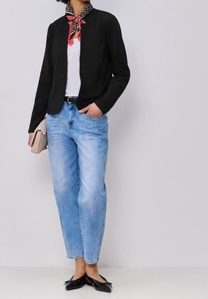 Person iført sort blazer, hvid top, lyseblå jeans, sorte flade sko og et mønstret rødt tørklæde, der holder en beige vævet clutchtaske.