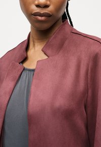 Κοντινό πλάνο ενός ατόμου που φοράει μπουφάν από καστόρι σε μωβ απόχρωση πάνω από γκρι μπλούζα, με εμφανές το λαιμό και το πάνω μέρος του στήθους και πλεξούδες στα μαλλιά.