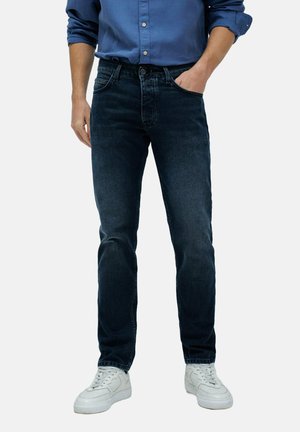Hombre vestido con jeans azul oscuro, camiseta de manga larga azul con las mangas arremangadas, zapatillas blancas, de pie con una mano en el bolsillo.