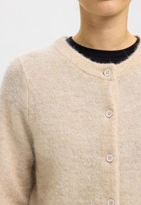 Ljusbeige kofta i mjuk ull med rund halsringning, sju knappar och en lätt luddig textur. Enkel och mysig design.