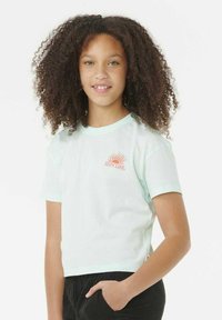 Chica con cabello rizado que lleva una camiseta corta azul clara con un sol y el logo "Rip Curl", y pantalones negros con una mano en el bolsillo.