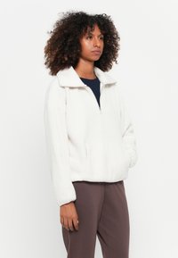 Vrouw met krullend haar, gekleed in een witte fleece jas en mauve broek, staand tegen een effen witte achtergrond.