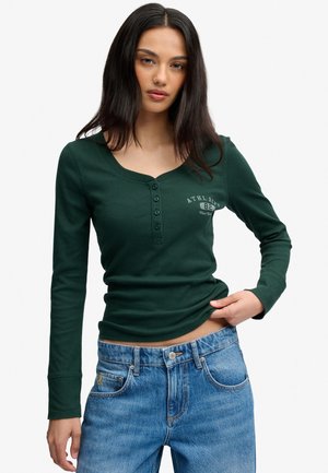Ung kvinde iført en mørkegrøn langærmet henley-shirt med knapper og blå jeans, der holder i skjortens kant med den ene hånd.
