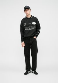 Giovane uomo in piedi con un cappellino nero, felpa nera con testo bianco, jeans neri e sneakers nere con righe bianche.