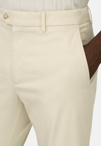 Gros plan sur un pantalon beige avec poche latérale, passants de ceinture et fermeture par bouton, une main à peau foncée partiellement dans la poche.