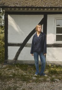 Laivaston kaksirivinen takki, jossa on suuret mustat napit, rento malli, yhdistettynä vaaleansinisiin farkkuihin; seisoo valkoisen olkikattoisen rakennuksen edessä.