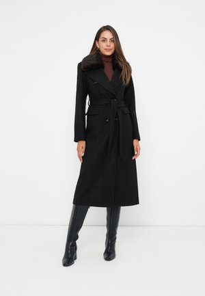 Mujer que lleva un abrigo largo negro con cinturón y cuello de piel, un jersey de cuello alto marrón y botas negras de tacón hasta la rodilla, de pie frente a un fondo blanco liso.