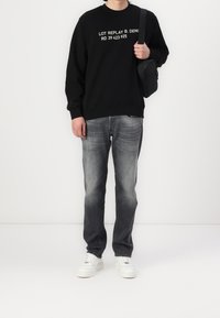 Felpa nera con testo bianco, jeans in denim grigio e sneakers bianche. L'outfit ha una vestibilità casual e un design semplice e moderno.