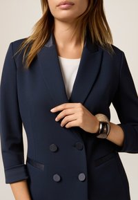 Marineblaues Doppelreiher-Blazer aus strukturiertem Stoff, mit Satinrevers und runden schwarzen Knöpfen. Modell trägt ein kreisförmiges Armband am Handgelenk.