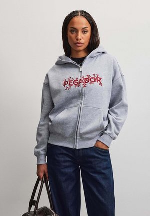 Ung kvinde med fletninger iført en lysegrå hættejakke med rødt blomstertryk og mørkeblå jeans, holder en brun lædertaske.