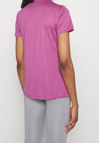 Osoba v krátkém rukávu fialové košile a světle šedých legínách, zachycena zezadu na čistém bílém pozadí.