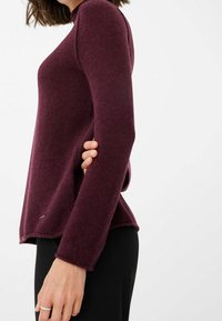 Pull bordeaux à manches longues, ourlet arrondi et texture douce. Présente un petit logo près du bord inférieur. Associé à un pantalon noir.