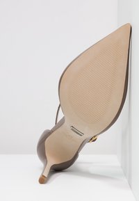 Chaussure à talon haut marron avec un bout pointu, un extérieur en cuir lisse et une semelle beige texturée. Présente une sangle décorative à la cheville.