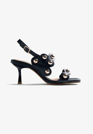 Sandalia de cuero negro con tira trasera, tacón de altura media, tiras con bordes ondulados y grandes tachuelas doradas brillantes como decoración.