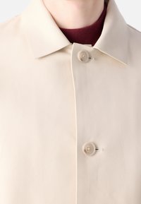 Gros plan d’un manteau beige à col avec deux boutons fermés sur un vêtement intérieur bordeaux à col montant.