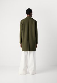 Olive green wool coat with a tailored fit, long sleeves, and a back vent, paired with wide white trousers, shown from the back.