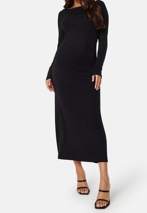 Kobieta w dopasowanej czarnej sukience midi z długim rękawem i okrągłym dekoltem oraz czarnych sandałach z odkrytymi palcami, stoi na tle jednolitego tła.