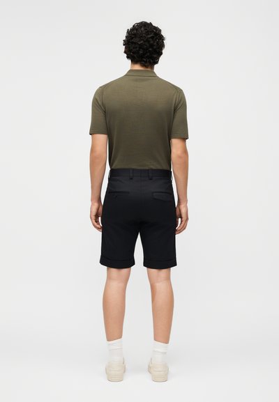 Άνδρας με σγουρά μαλλιά που φοράει πράσινο λαδί κοντομάνικο πόλο, μαύρα σορτς με μανσέτα, λευκές κάλτσες και λευκά αθλητικά παπούτσια, στέκεται με την πλάτη προς τα εμπρός.