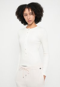 Top cu mâneci lungi de culoare crem, cu decolteu rotund și nasturi. Are o textură nervurată și se potrivește strâns corpului.