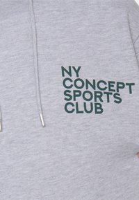 Huppari, joka on valmistettu pehmeästä puuvillasekoitteesta, ja siinä on suuri vihreä printti "NY CONCEPT SPORTS CLUB" edessä. Hopeiset metalliset päätypalat nyöreissä.