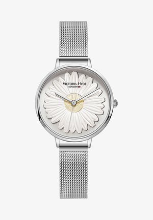 Reloj con correa de malla plateada que presenta un diseño floral blanco en la esfera, con pétalos en relieve y un detalle dorado en el centro; nombre de la marca visible.