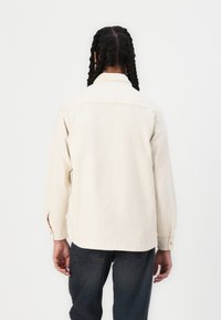 Vaalean beige paita, joka on valmistettu paksusta puuvillasta, pitkät hihat ja viiston kauluksen kanssa. Varustettu nappikauluksilla ja suoran helman kanssa. Yhdistetty tummiin denimpaitoihin.