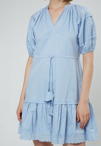 Hellblaues Baumwollkleid mit V-Ausschnitt, Puffärmeln und Spitzenbesatz. Es hat eine Taillenbindung und einen mehrlagigen Volantabschluss mit Lochmuster.