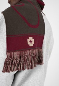 Strikket tørklæde i mørkebrun og burgunder med et beige medicinsk korsdesign og frynse detaljer; blød tekstur og rektangulær form.