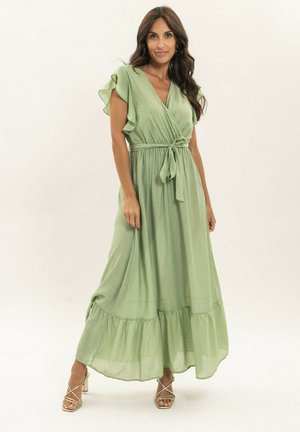 Femme aux longs cheveux bruns portant une robe portefeuille vert clair à manches volantées et des talons beiges à brides, debout devant un fond uni.