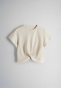 Beżowy krótki top z krótkim rękawem, okrągłym dekoltem i skręconym węzłem na środku z przodu, prezentowany na jasnym jednolitym tle.