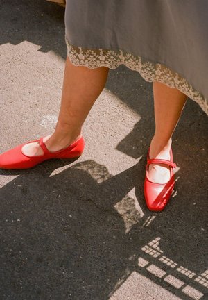 Scarpe Mary Jane in pelle rossa, con punta arrotondata, tacco piatto e cinturino alla caviglia, posizionate su una superficie grigia testurizzata con orlo di pizzo.