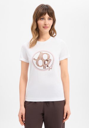 Frau trägt ein weißes T-Shirt mit kurzen Ärmeln und einem großen "JOOP!"-Logo in Beige und Braun, kombiniert mit dunkelbrauner Hose.