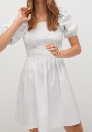 Femme portant une robe blanche à manches bouffantes et un corsage smocké, debout devant un fond clair uni.