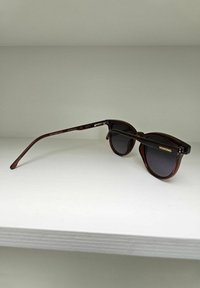 Lunettes de soleil en écaille marron avec des verres foncés, de forme ronde et à branches fines. Présente le logo "KOMONO" sur la branche et offre une protection UV.