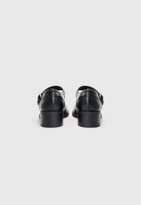 Black leather shoes with a low block heel, featuring embossed detailing and an adjustable buckle on the strap. Smooth texture.