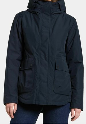 Femme portant une veste imperméable bleu marine avec un col montant, des poches à rabat à l'avant avec boutons-pression, et une fermeture éclair.