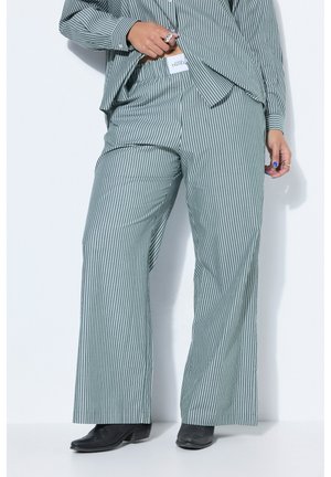 Persona che indossa pantaloni a righe verticali grigi e bianchi a gamba larga e camicia coordinata, tenendo la camicia sollevata per mostrare l'etichetta della vita.