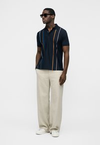 Man wearing dark polo shirt with vertical stripes, beige wide-leg pants, white sneakers, and sunglasses, standing with hands in pockets.