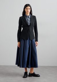 Svart kavaj med två framfickor, lager på en mönstrad skjorta. Flödande marinblå kjol och svarta snörskor fulländar outfiten.