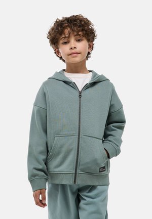 Garçon aux cheveux bouclés portant un sweat à capuche zippé vert clair et un pantalon assorti, debout avec une main dans la poche devant un fond blanc.